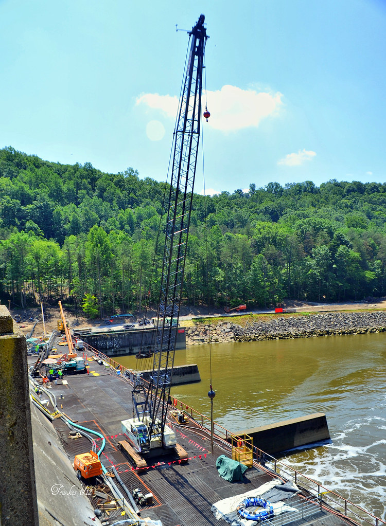 Crane on a floating platform at a river dock with construction equipment and workers, backed by a forested shore.